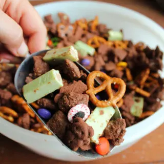 Hand holding a scoop filled with Halloween snack mix made from chocolaty puppy chow, pretzels, teddy grahams, Halloween M&M's, green kit kats, and candy eyeballs.