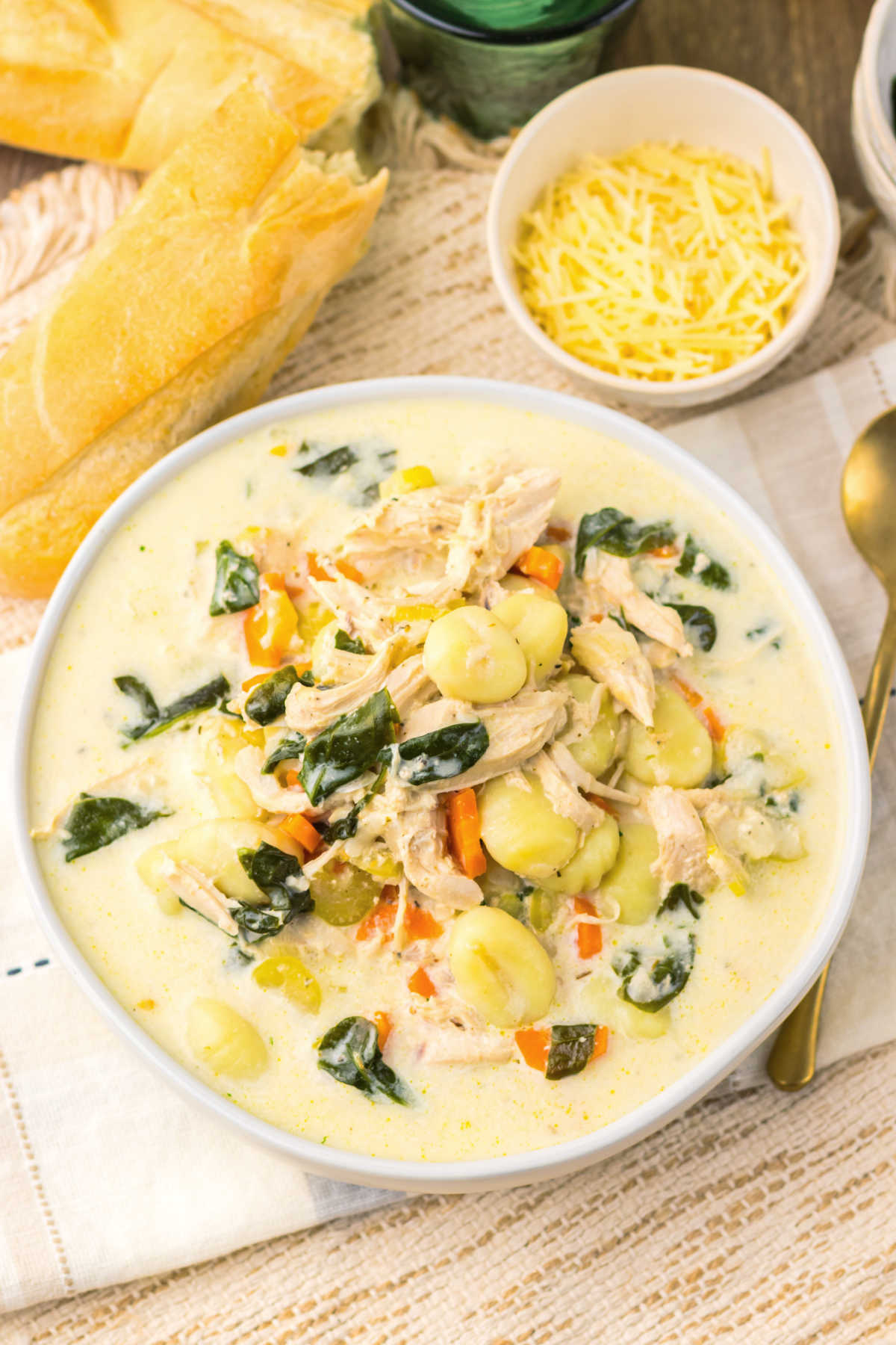 Bowl of slow cooked chicken gnocchi soup with shredded chicken, gnocchi, spinach, and veggies in a creamy broth next to a bowl of shredded parmesan cheese and a baguette, ready to eat. 