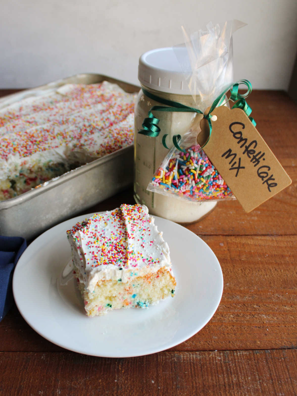 Piece of confetti cake with homemade condensed milk frosting and sprinkles on top next to a jar of homemade cake mix with sprinkles. 