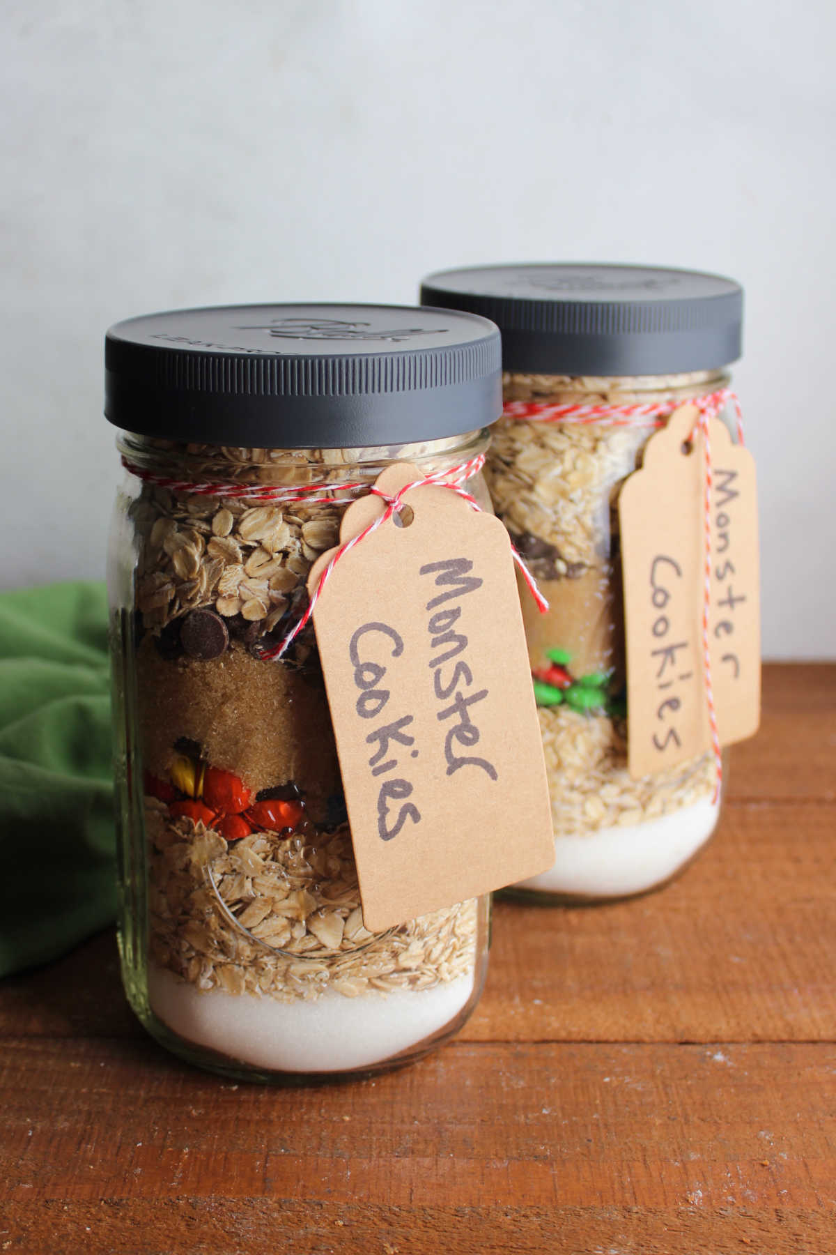 Jars of monster cookie mix with the dry ingredients in layers inside the jars and tags tied to the outside with baking instructions. 