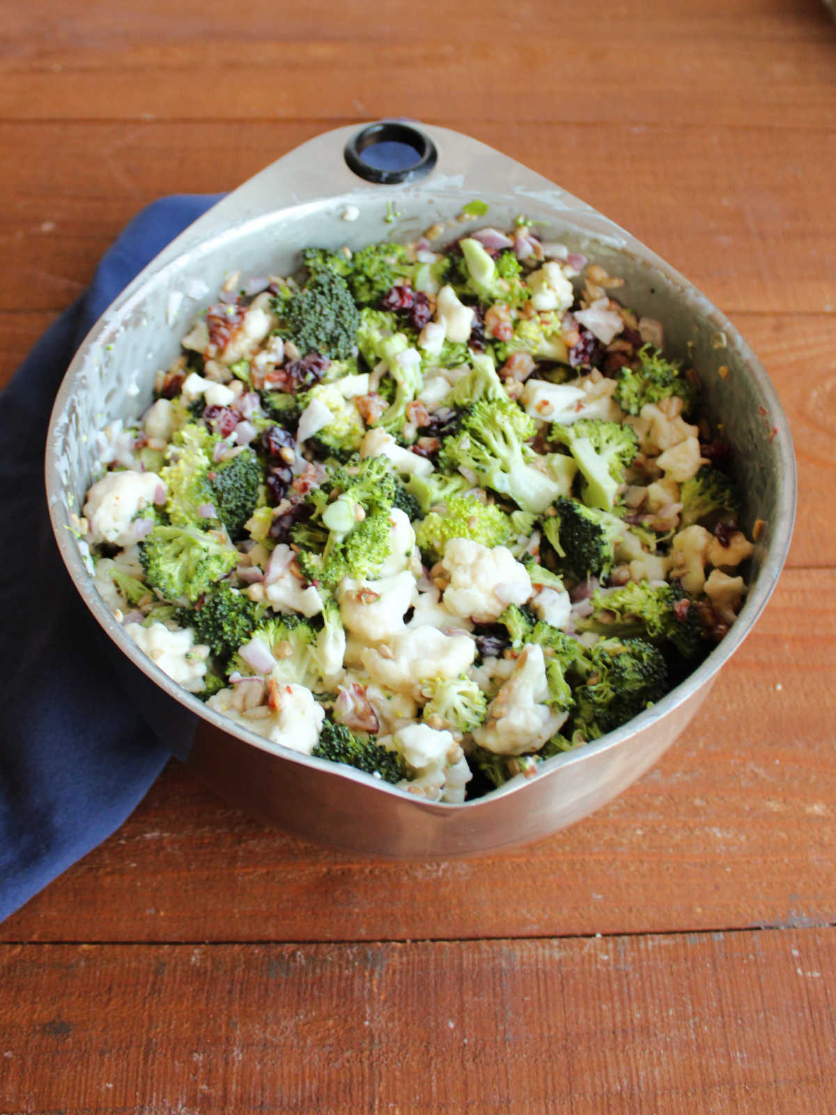 Large mixing bowl filled with broccoli salad with bits of broccoli, cauliflower and red onion with sunflower seeds, bacon, and craisins in light dressing. 