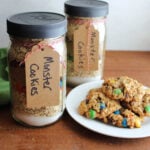 Two jars of layered monster cookie mix next to a plate of freshly baked monster cookies.