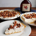 Slice of no-bake pecan cream pie with creamy maple filling topped with chopped pecans next to two pecan cream pies and a bottle of maple syrup.