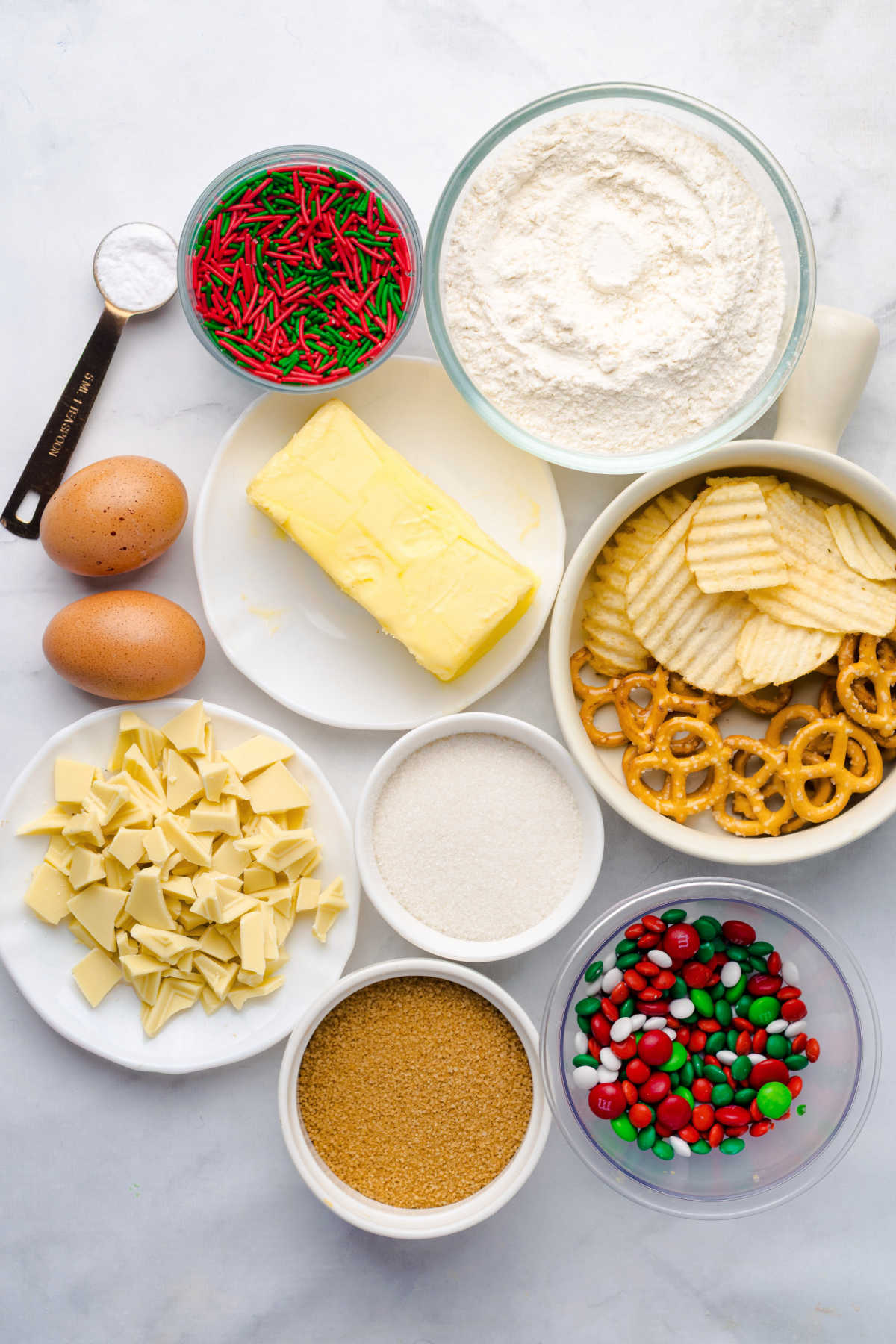 Ingredients including butter, flour, sugar, brown sugar, eggs, vanilla, pretzels, potato chips, sprinkles, and candies ready to be made into Christmas kitchen sink cookies. 