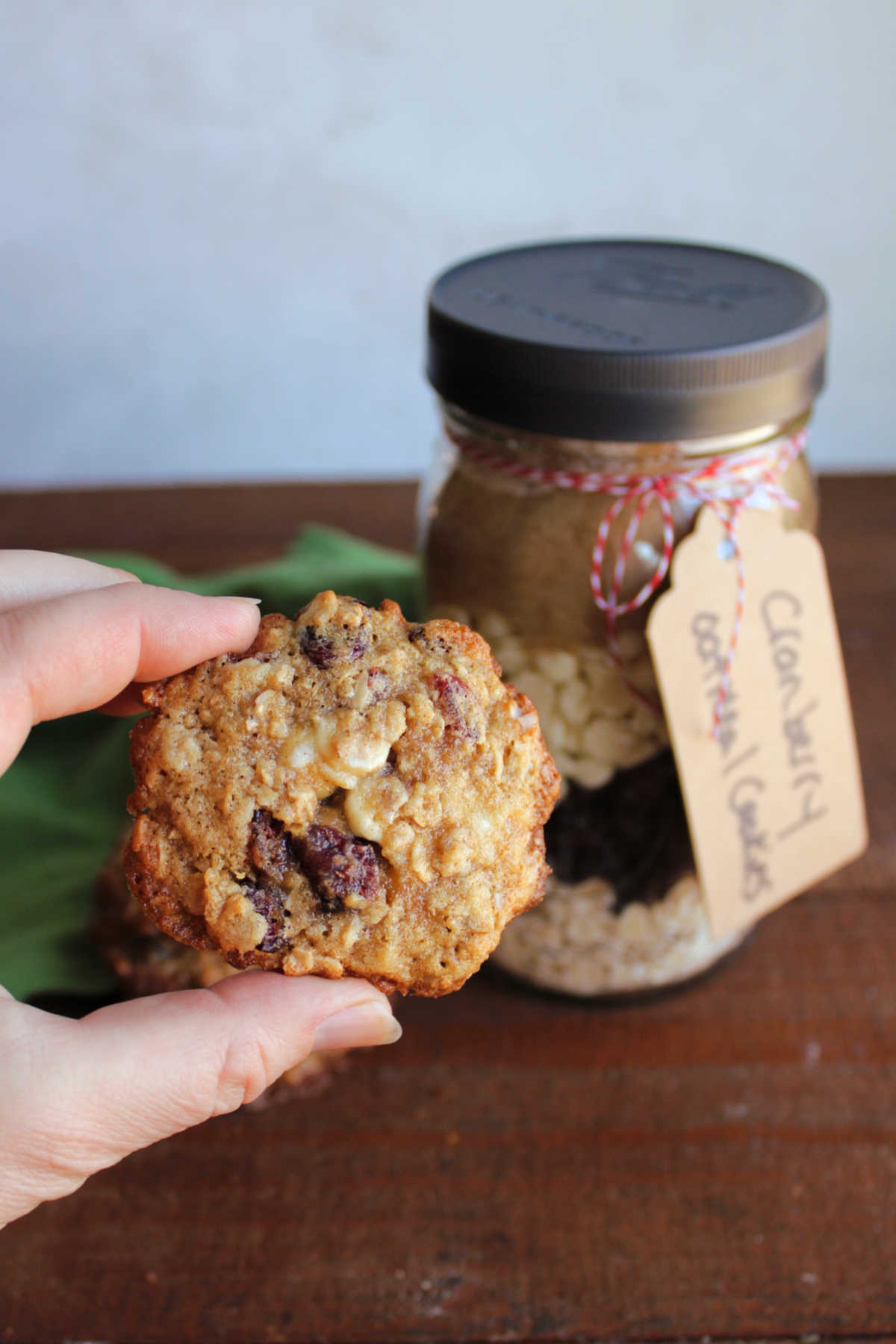 Hand holding a cranberry white chocolate oatmeal cookie next to a jar of homemade cookie mix. 