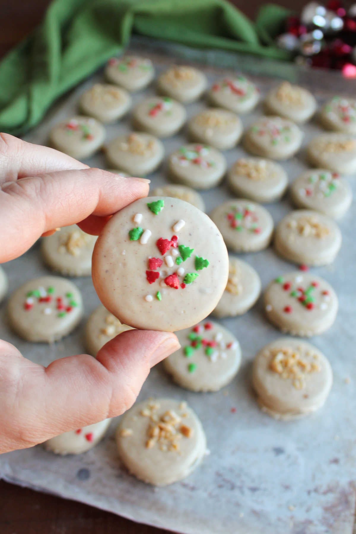 Hand holding peanut butter dipped Oreo with Christmas sprinkles with wax paper lined tray of more dipped Oreos in the background. 