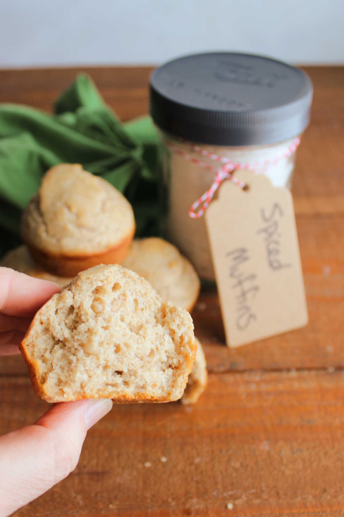 Hand holding a half of a spiced muffin showing the soft texture inside with a jar of muffin mix and more baked muffins in the background. 