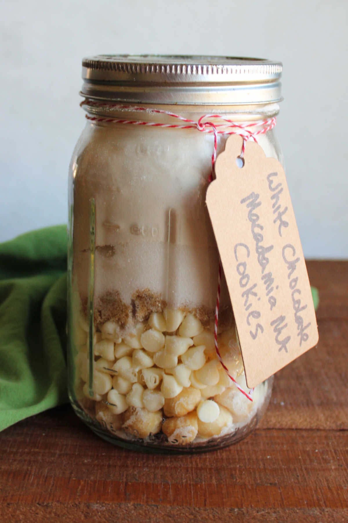 Quart jar filled with layers of macadamia nuts, white chocolate chips, brown sugar, white sugar, and flour mixed with salt, baking powder and baking soda to make white chocolate macadamia nut cookies. 