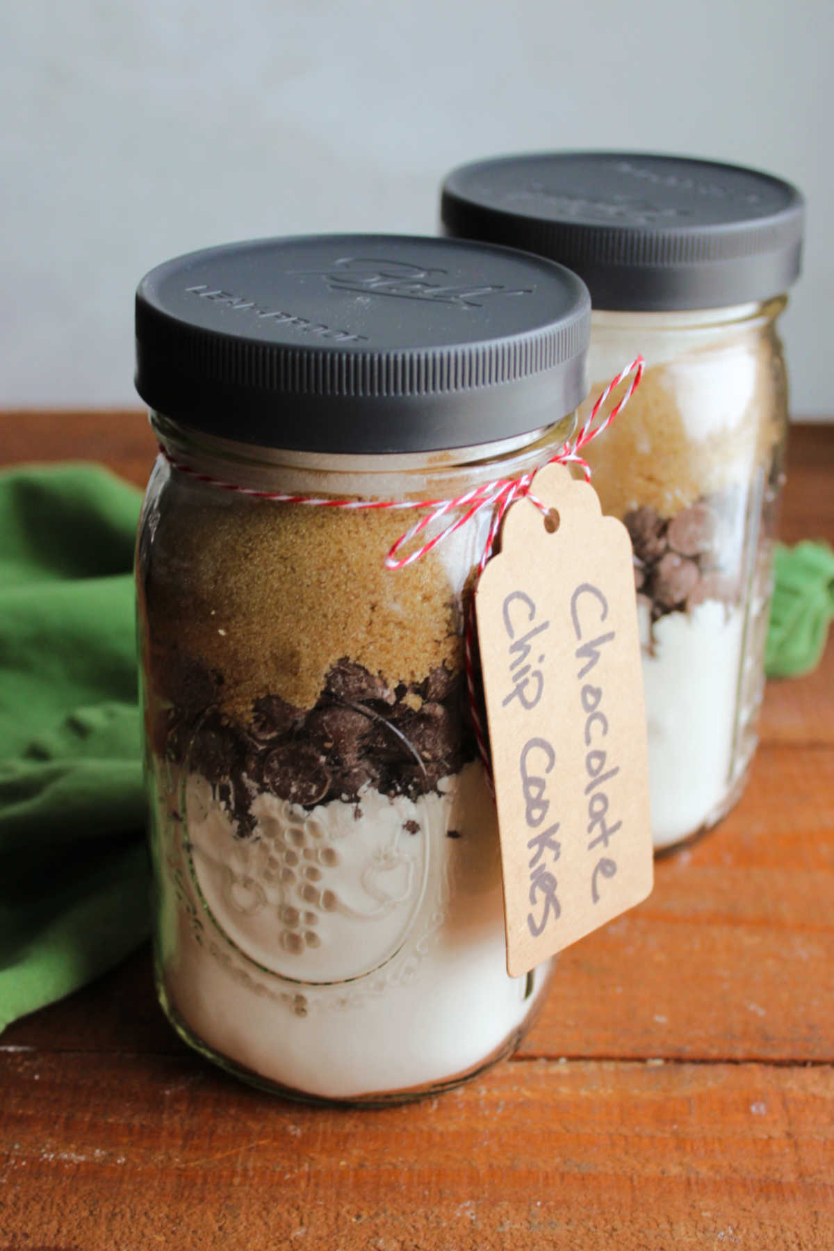 Two glass quart jars filled with the dry ingredients to make chocolate chip cookies including a flour mixture, chocolate chips, brown sugar, and granulated sugar with a tag tied to the jar. 