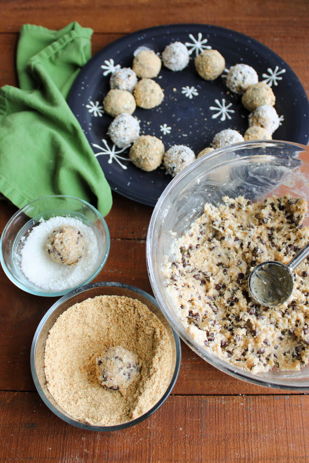 Bowl of chilled moose fart mixture with small cookie scoop to transfer the candy into the nearby bowl of graham cracker crumbs or shredded coconut with some formed moose fart balls on a plate in the background. 