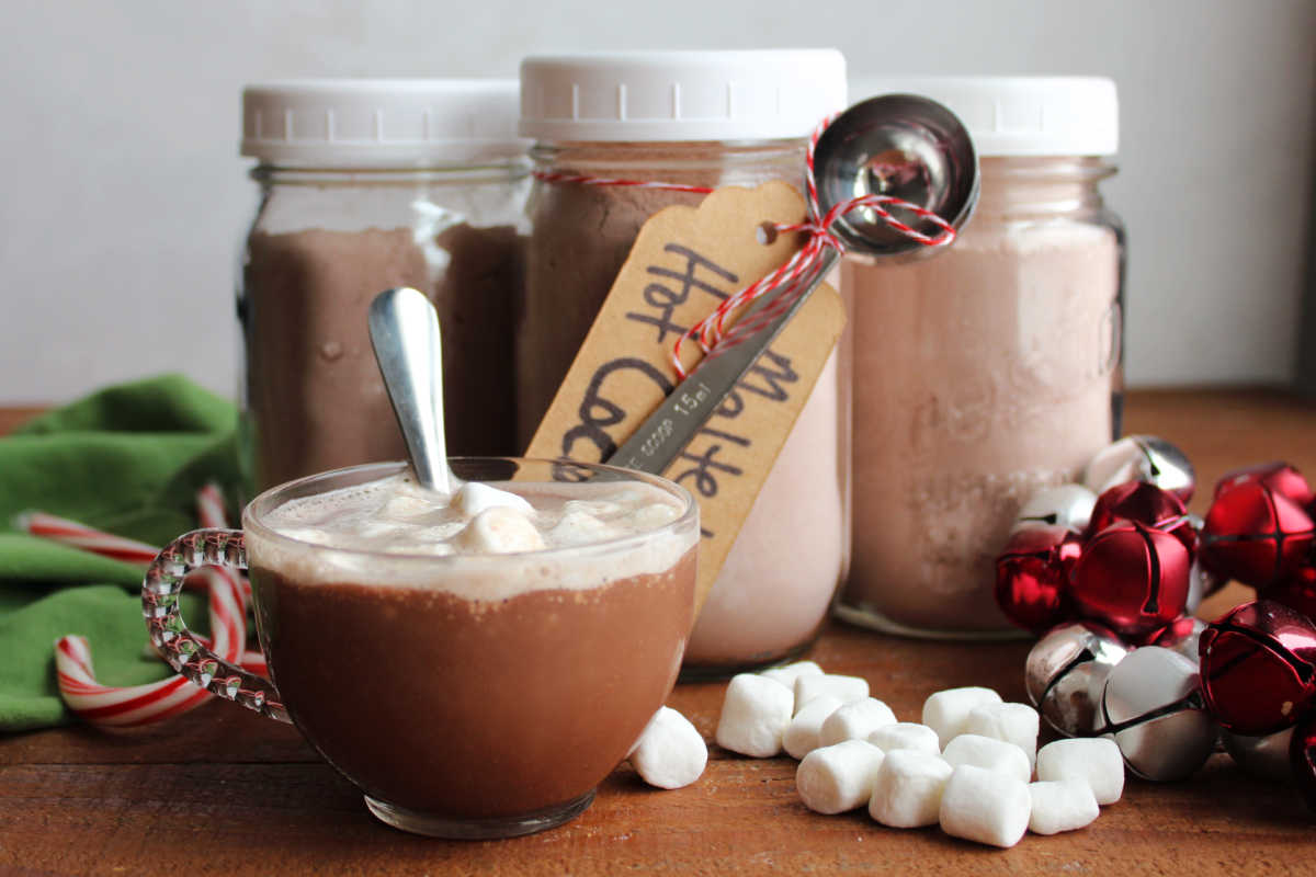 Mug of malted hot chocolate with marshmallows next to 3 pint jars of homemade hot chocolate mix. 
