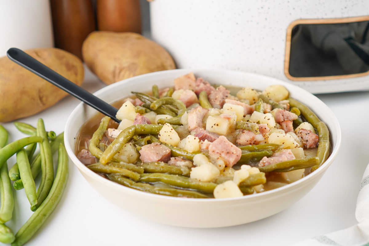 Crockpot Green Beans With Ham and Potatoes 5 Bowl of green beans with chunks of ham and potatoes in them in front of the slow cooker that they were cooked in.