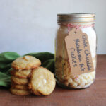 Jar of homemade white chocolate macadamia nut cookie mix next to a stack of buttery homemade cookies.