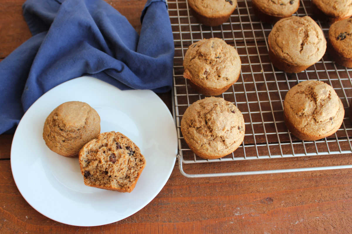 Two homemade bran muffins on a plate with a wire cooling rack of more bran muffins nearby. 