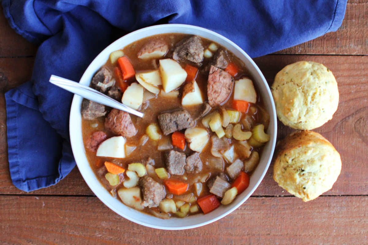 Bowl of venison stew with chunks of tender meat and veggies in thick broth with a couple of savory muffins, ready to eat. 