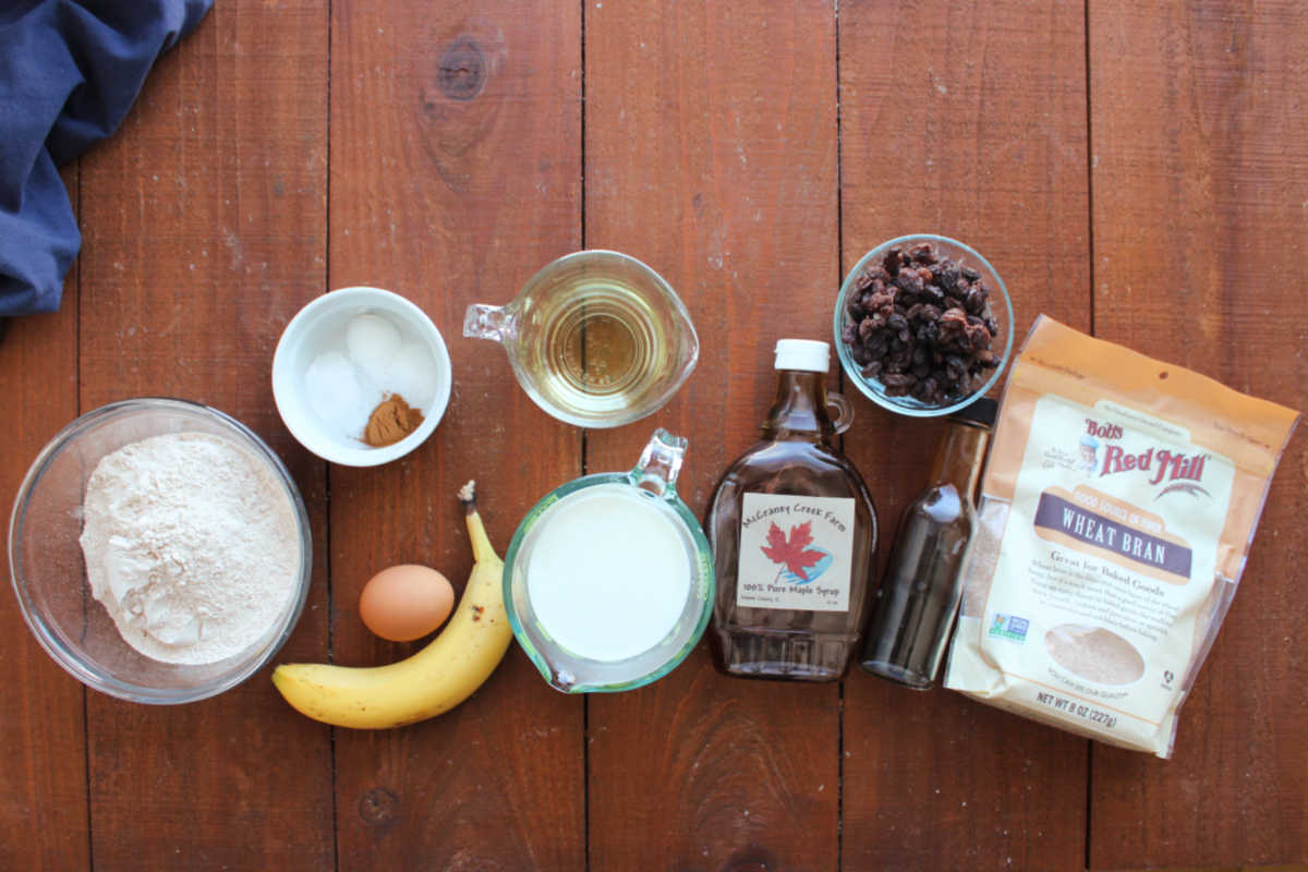 Ingredients including whole wheat flour, baking powder, baking soda, salt, cinnamon, egg, banana, milk, oil, maple syrup, raisins, vanilla and wheat bran ready to be made into bran muffins. 
