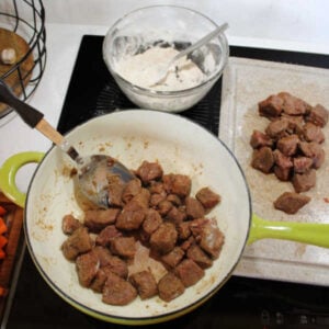 Browning coated venison in batches with some browned meat on a cutting board, some in the pot, and the bowl of extra flour nearby.