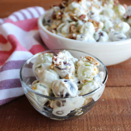 Small glass bowl of creamy grape salad with red and green grapes coated in cream dressing with bits of brown sugar and chopped pecans on top with a serving bowl of more grape salad in the background.