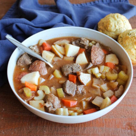 Bowl of venison stew with chunks of meat, potatoes, carrots, onions and celery in rich broth with some savory parmesan herb muffins nearby.