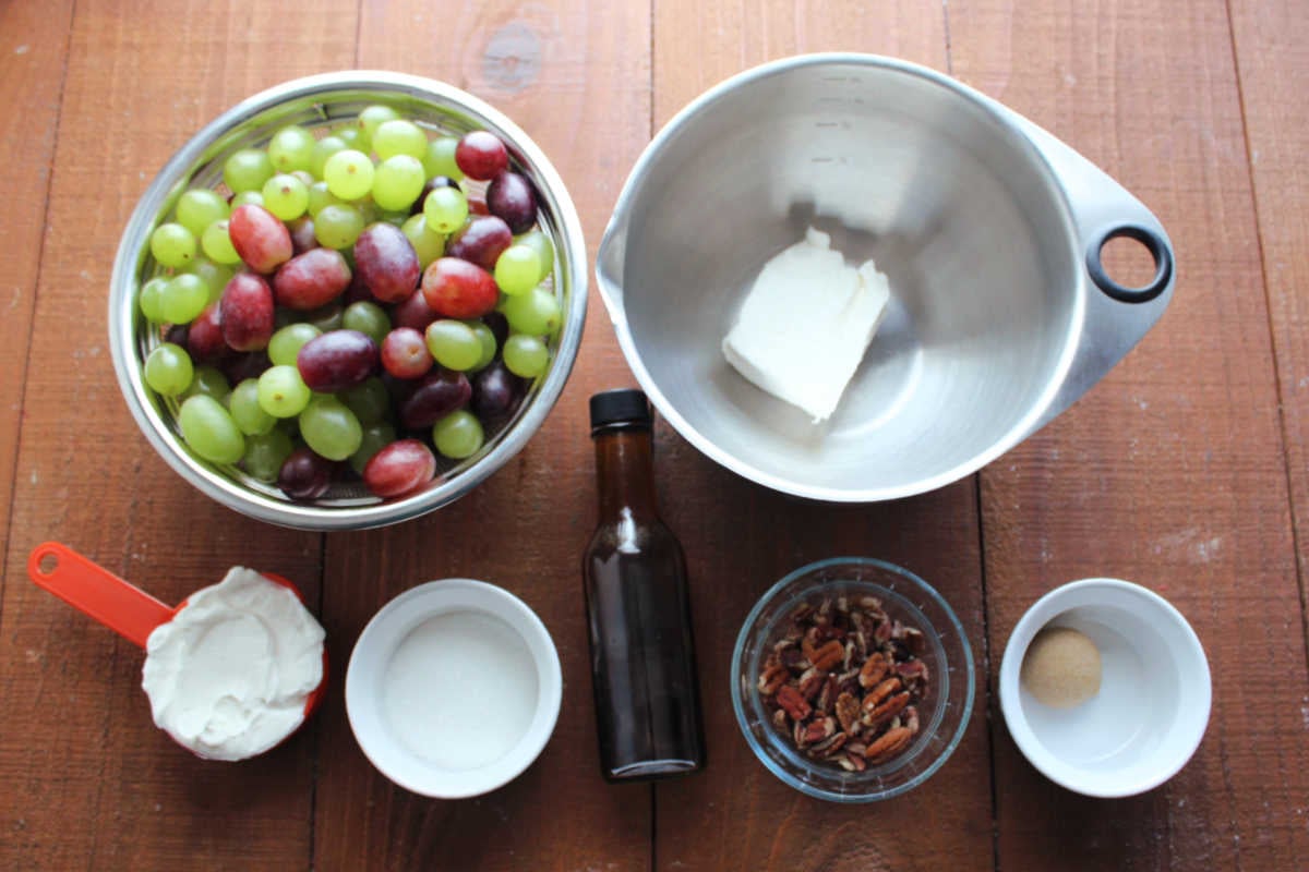 Ingredients including red and green seedless grapes, cream cheese, sour cream, sugar, vanilla, pecans, and brown sugar ready to be made into a grape salad. 