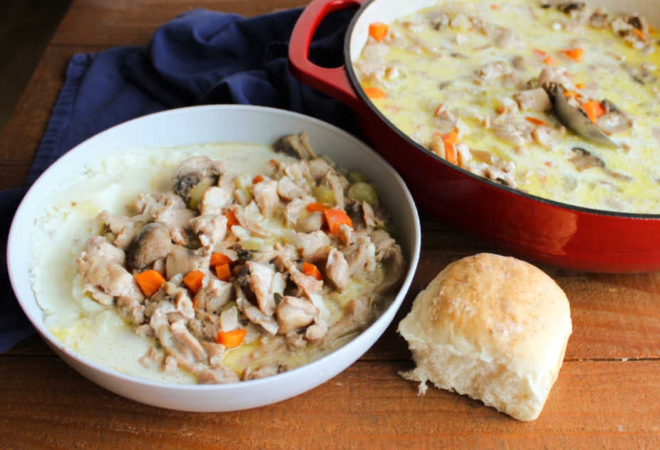 Bowl of rabbit stew with a creamy broth and chunks of veggies and mushrooms served over mashed potatoes with a homemade dinner roll nearby. 