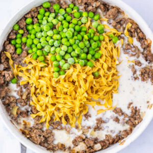 Amish Country Casserole 12 Skillet filled with ground beef and onion in sauce made from milk and cream of mushroom soup with uncooked egg noodles and peas on top.