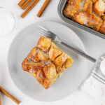 square of donut bread pudding on dessert plate with baking pan with more bread pudding and some cinnamon sticks nearby.