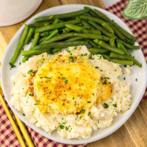 Dinner plate with green beans and a pile of mashed potatoes topped with a chicken breast covered in melted cheese, a parmesan ranch mixture and golden brown bread crumbs, ready to eat.