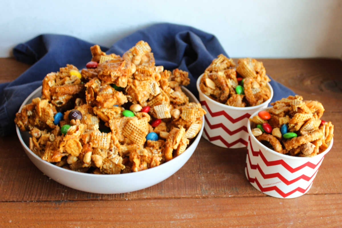 Peanut Butter Chex Mix 6 Serving bowl piled high with crunchy peanut butter Chex mix next to two paper cups filled with more Chex mix, ready to eat.