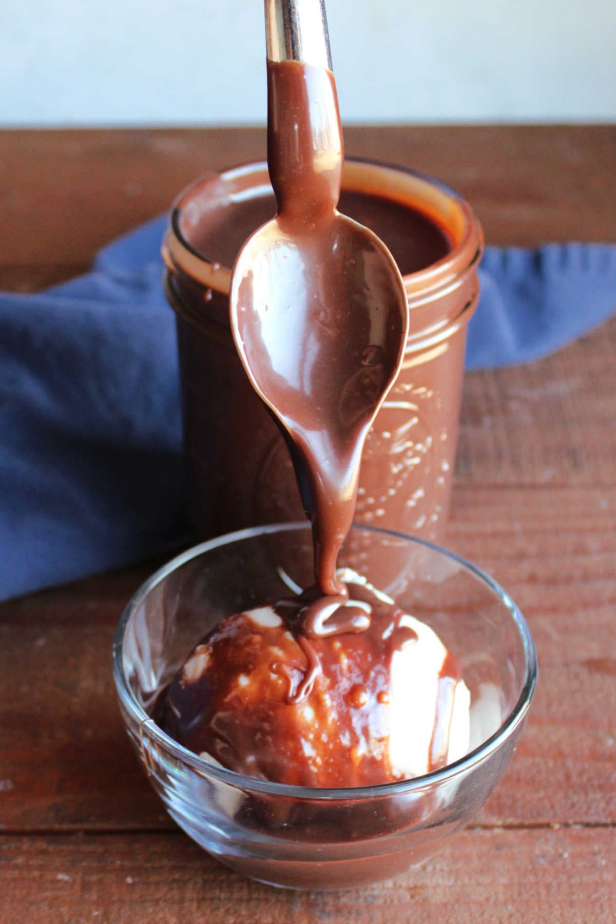 Spoon drizzling warm hot fudge sauce over a bowl of ice cream. 