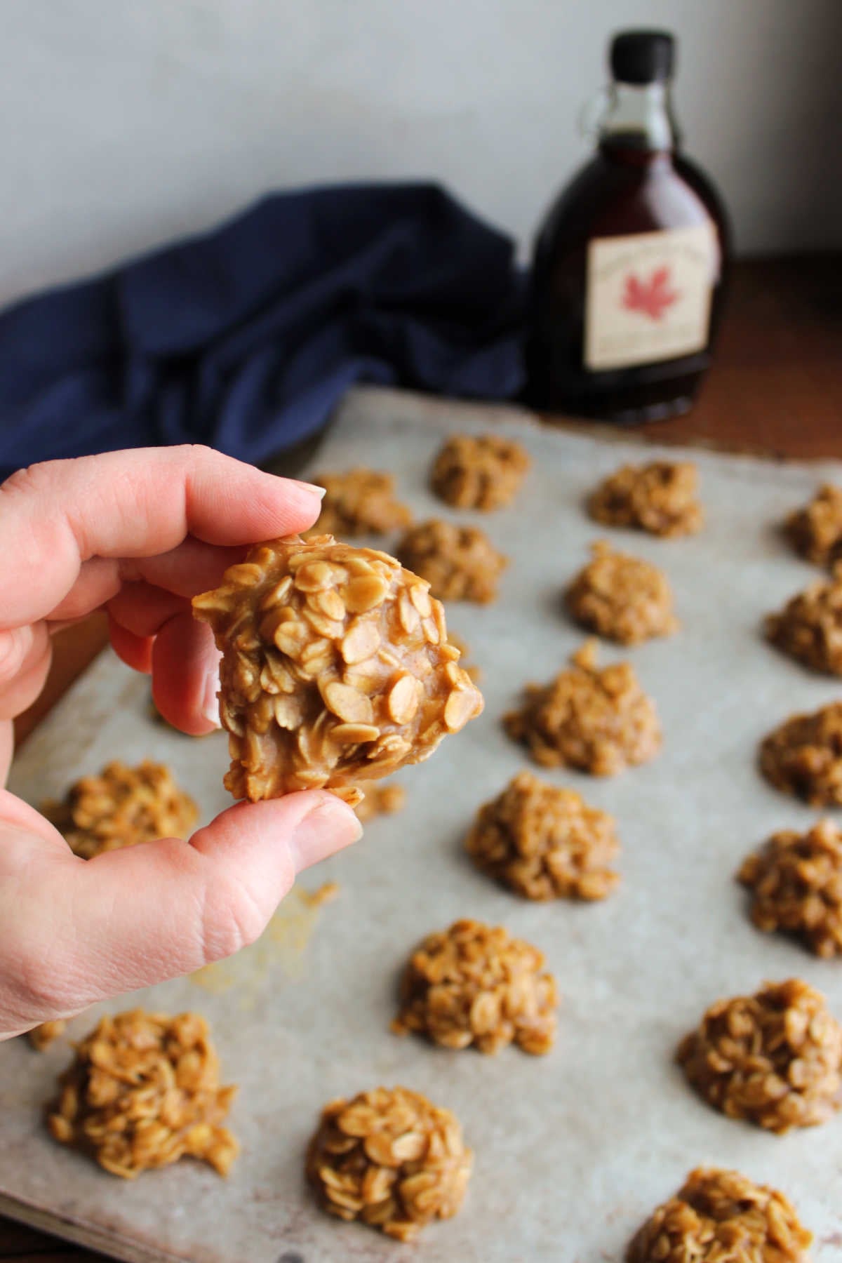 Hand holding a no bake maple cookie over a tray with more cookies that have been scooped on wax paper to set. 
