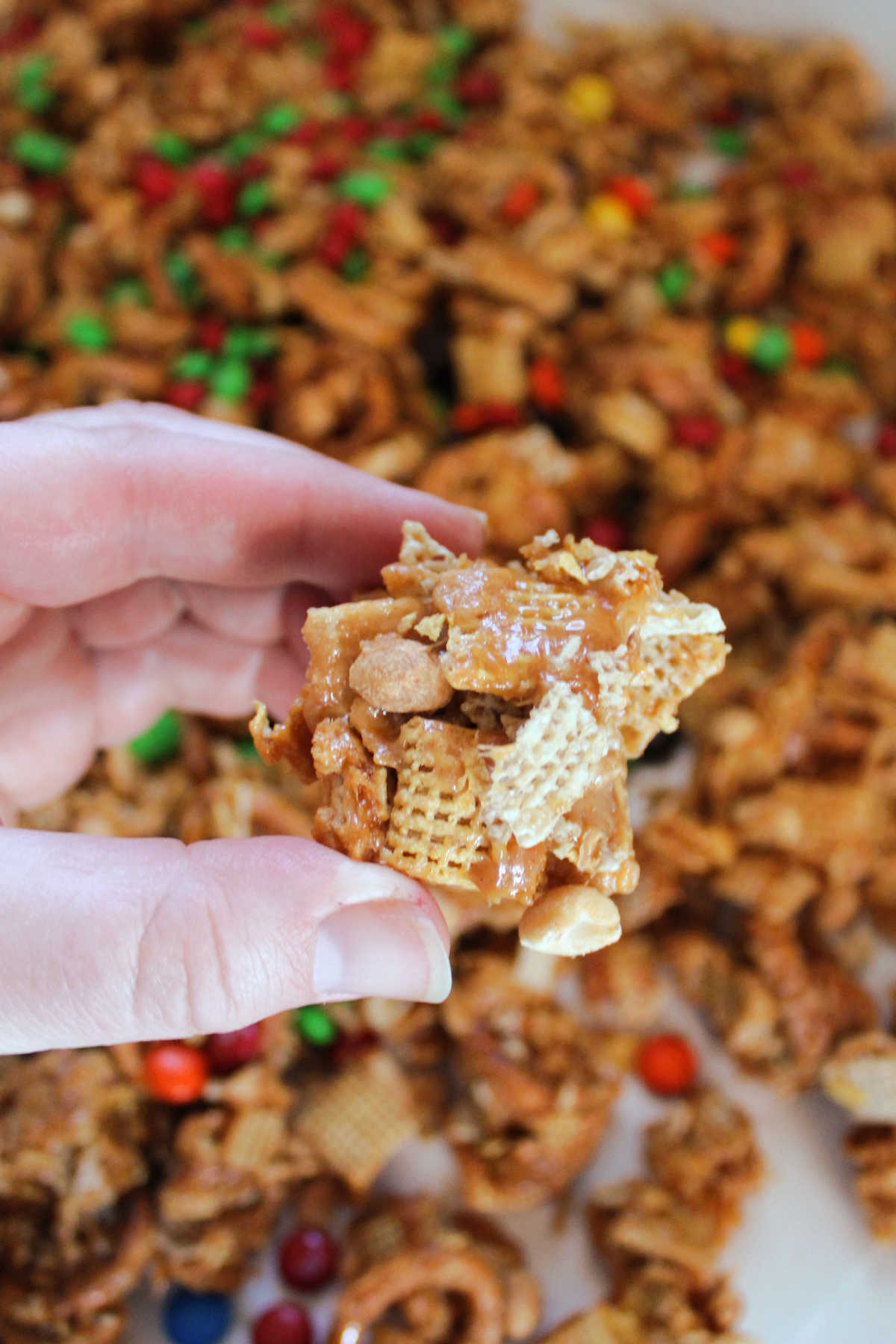 Peanut Butter Chex Mix 8 Hand holding a cluster of Chex cereal, corn flakes, pretzels, and peanuts coated in peanut butter caramel over the rest of the snack mix cooling on sheets of wax paper.