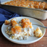 Plate filled with a piece of breakfast casserole with a layer of biscuits topped with scrambled eggs, creamy sausage gravy, tater tots and melted cheese, ready to eat.