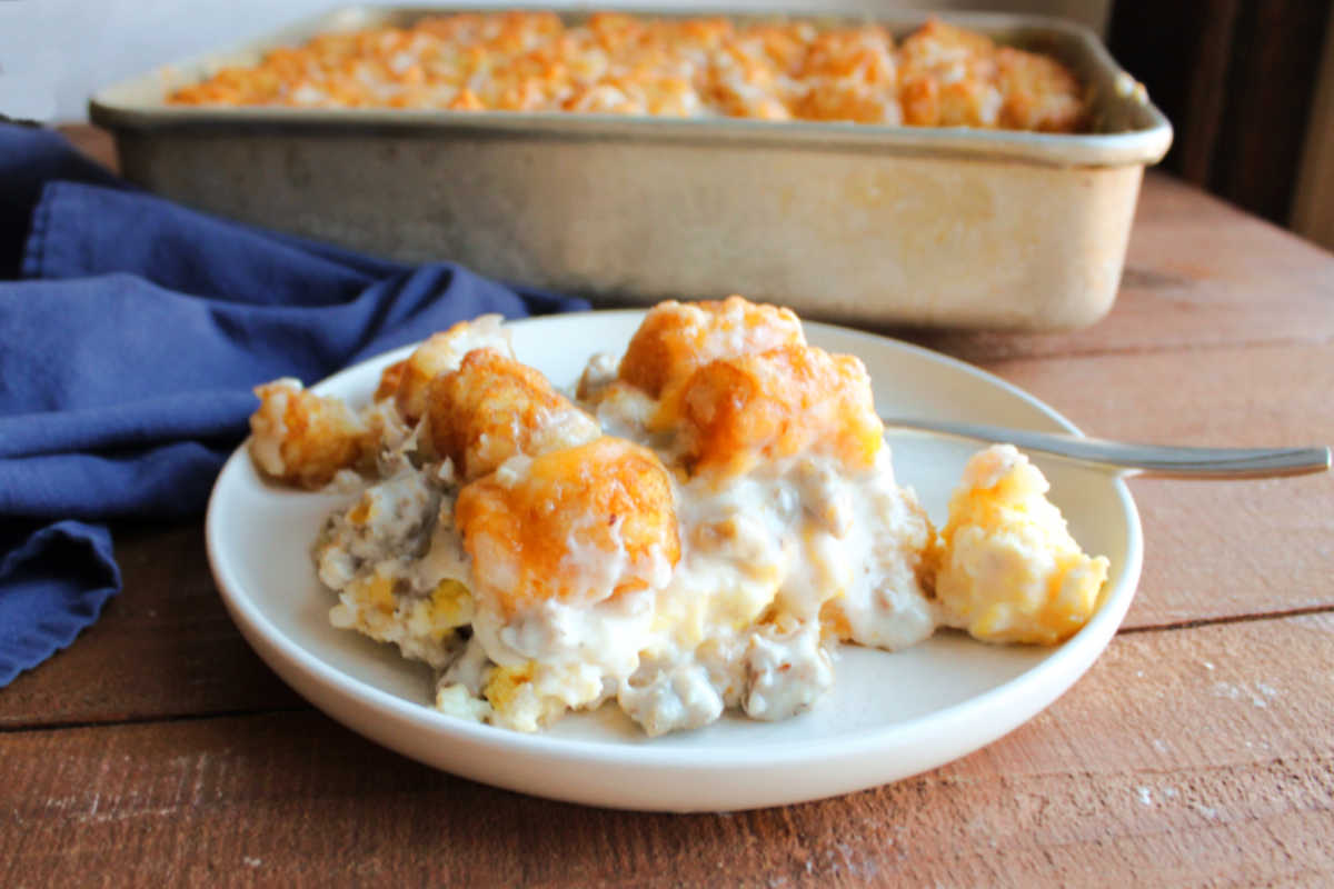 Biscuits and Gravy Tater Tot Casserole 5 Plate with a piece of sausage gravy tater tot casserole on it showing the layers of biscuits, eggs, gravy, and tater tots with a fork and the pan of remaining casserole in the background.