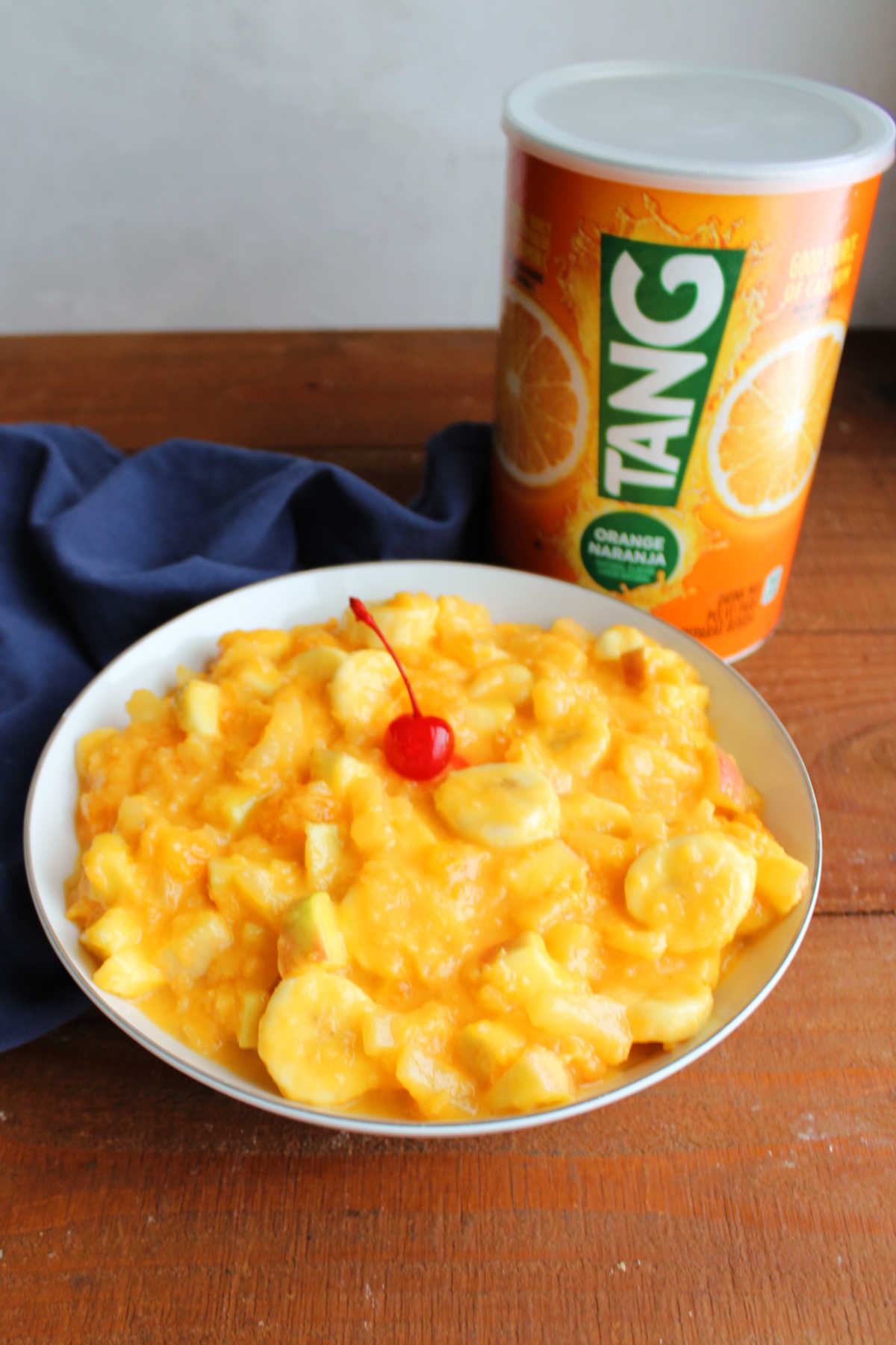 Tang Fruit Salad 8 Old fashioned china serving bowl filled with Tang fruit salad topped with a maraschino cherry next to a large container of Tang powder.