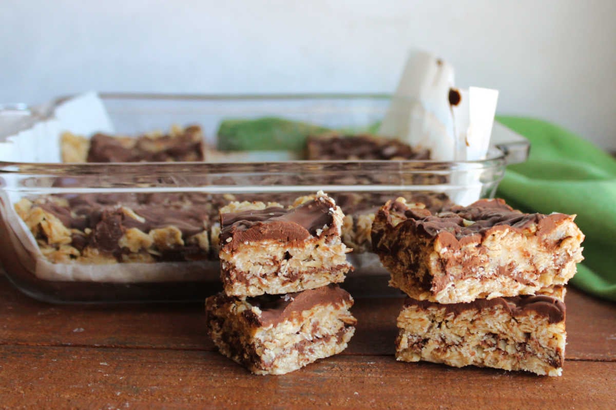 Small squares of no bake oatmeal cookie bars with chocolate and peanut butter filling in front of a pan with more bars inside. 