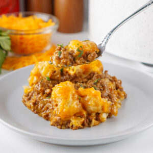 Fork with a bite of taco tater tot casserole on it showing cheesy seasoned beef mixture with golden tater tots with melted cheese on top.