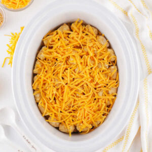 Tater tots and shredded cheese over the top of the cheesy taco meat mixture in a crockpot, ready to cook.