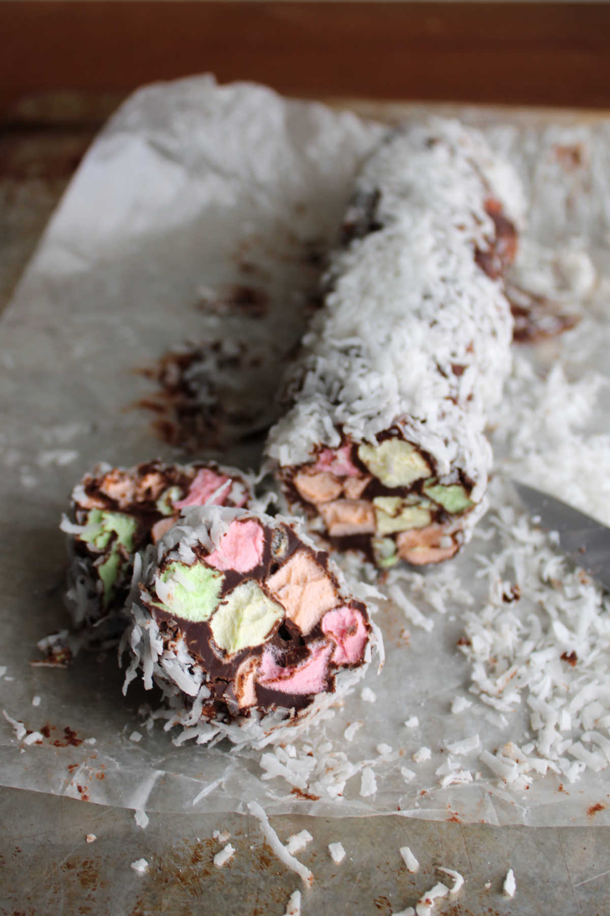 Knife cutting a coconut coated log of stained glass candy into individual "cookies" making little chocolate rounds with bits of fruity marshmallow inside. 