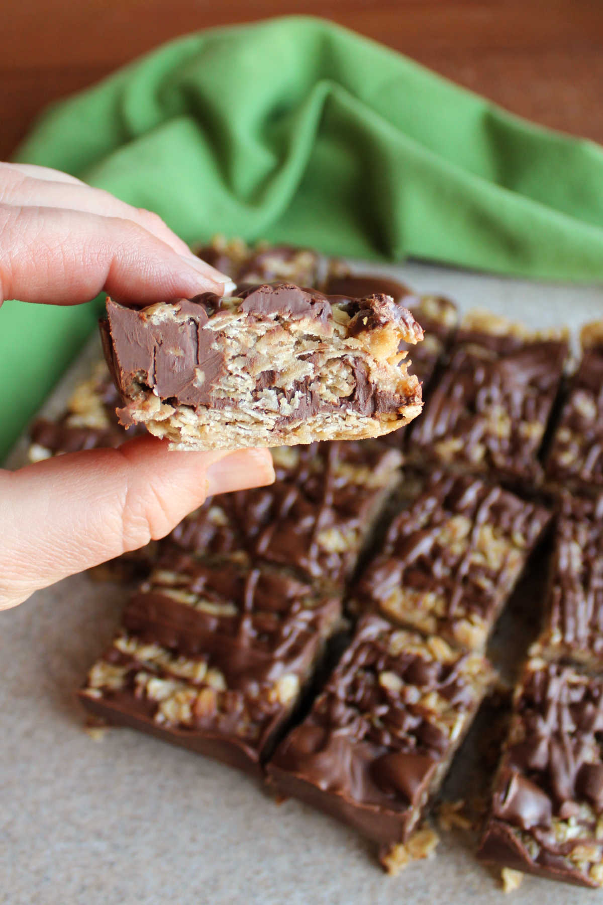 Hand holding a no bake oatmeal cookie bar with a chocolate layer in the middle and on the top with a cutting board with more bars in the background. 