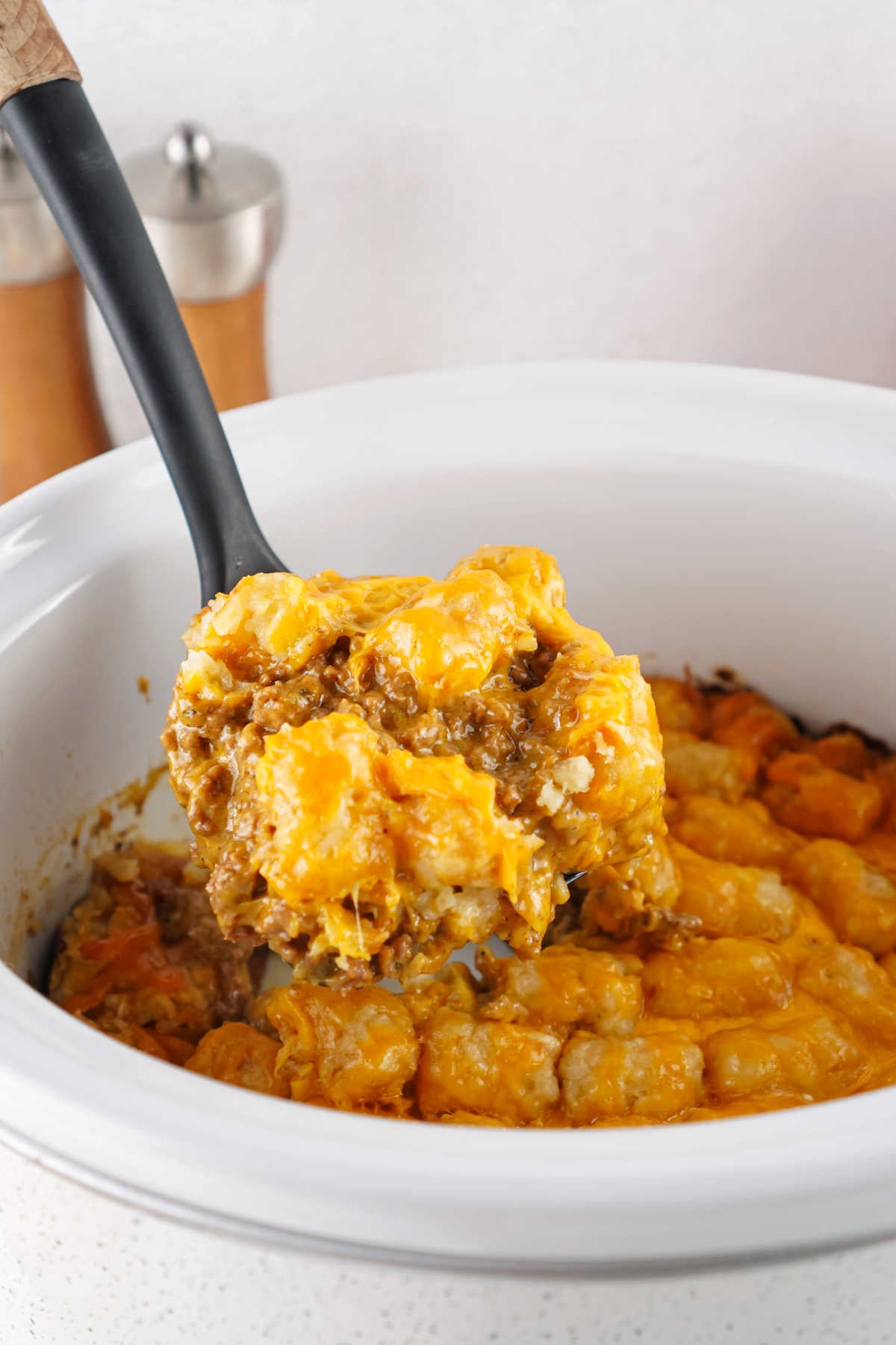 Serving spoon getting a helping of taco tater tot casserole out of a crockpot showing cheesy seasoned ground beef topped with tater tots and melted cheese. 