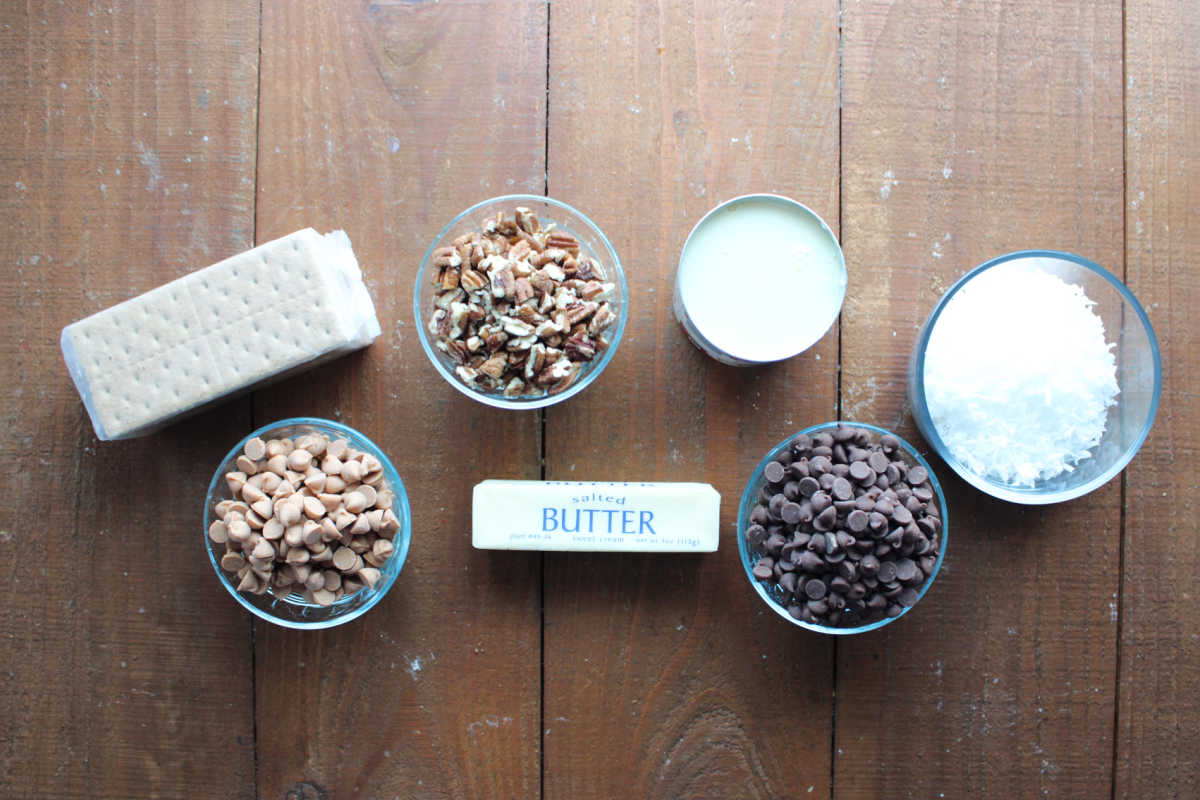 Ingredients including graham crackers, chopped pecans, sweetened condensed milk, flaked coconut, chocolate chips, butter and butterscotch chips ready to be made into 7 layer bars. 