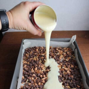 Hand drizzling a can of sweetened condensed milk over the pan with the coconut and chocolate chips etc.