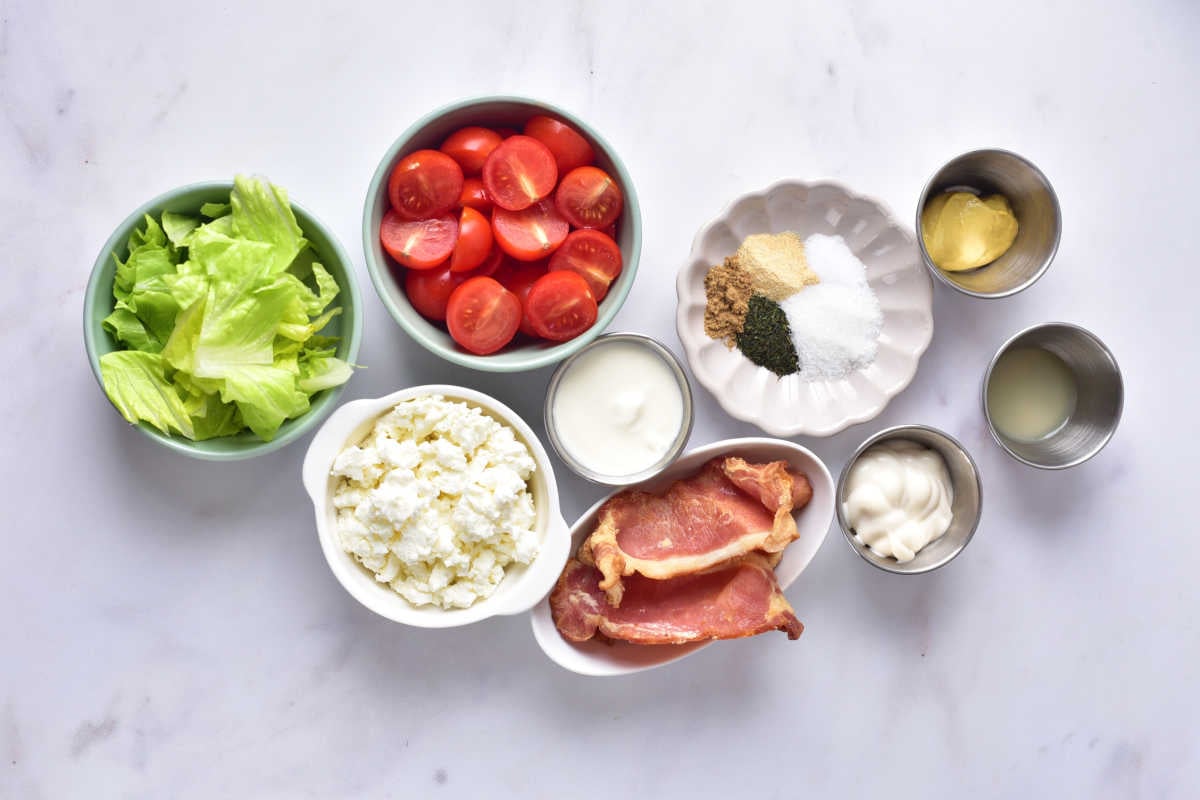 Ingredients including lettuce, cherry tomato halves, feta cheese, bacon, mayonnaise, sour cream, mustard, lemon juice, and seasonings ready to be made into a blt salad. 