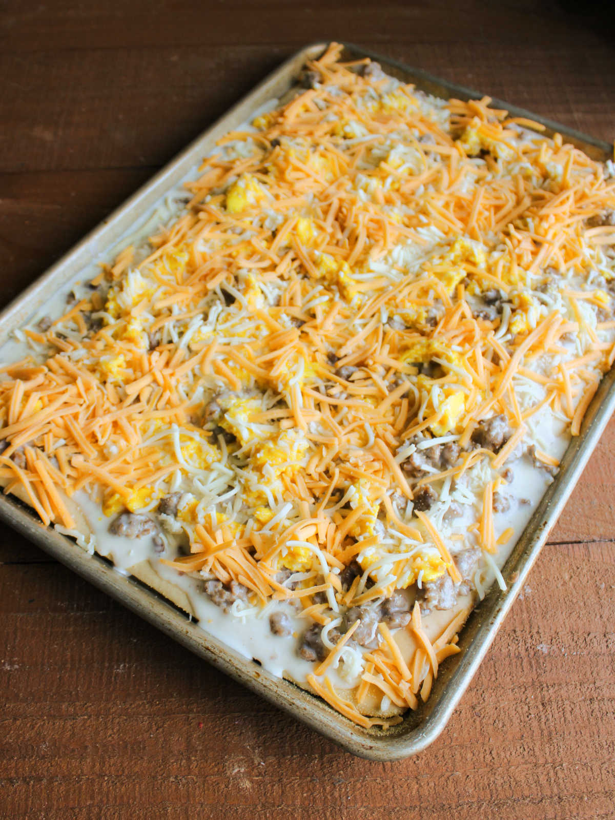 Sheet pan filled with partially baked pizza crust topped with sausage gravy, soft scrambled eggs, and cheese, ready to go into the oven to bake. 