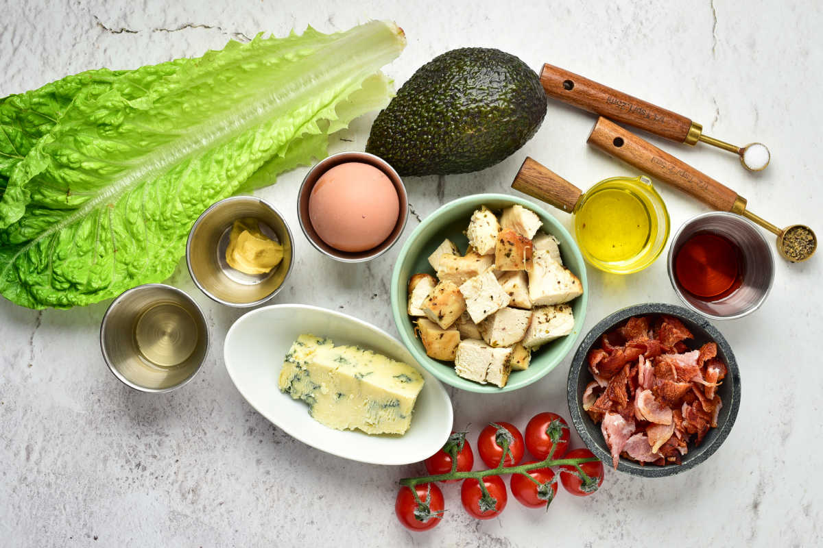 Ingredients including romaine lettuce, avocado, eggs, blue cheese, bacon, chicken breast tomatoes, mustard, olive oil, vinegar, salt and pepper, ready to be made into a homemade cobb salad. 