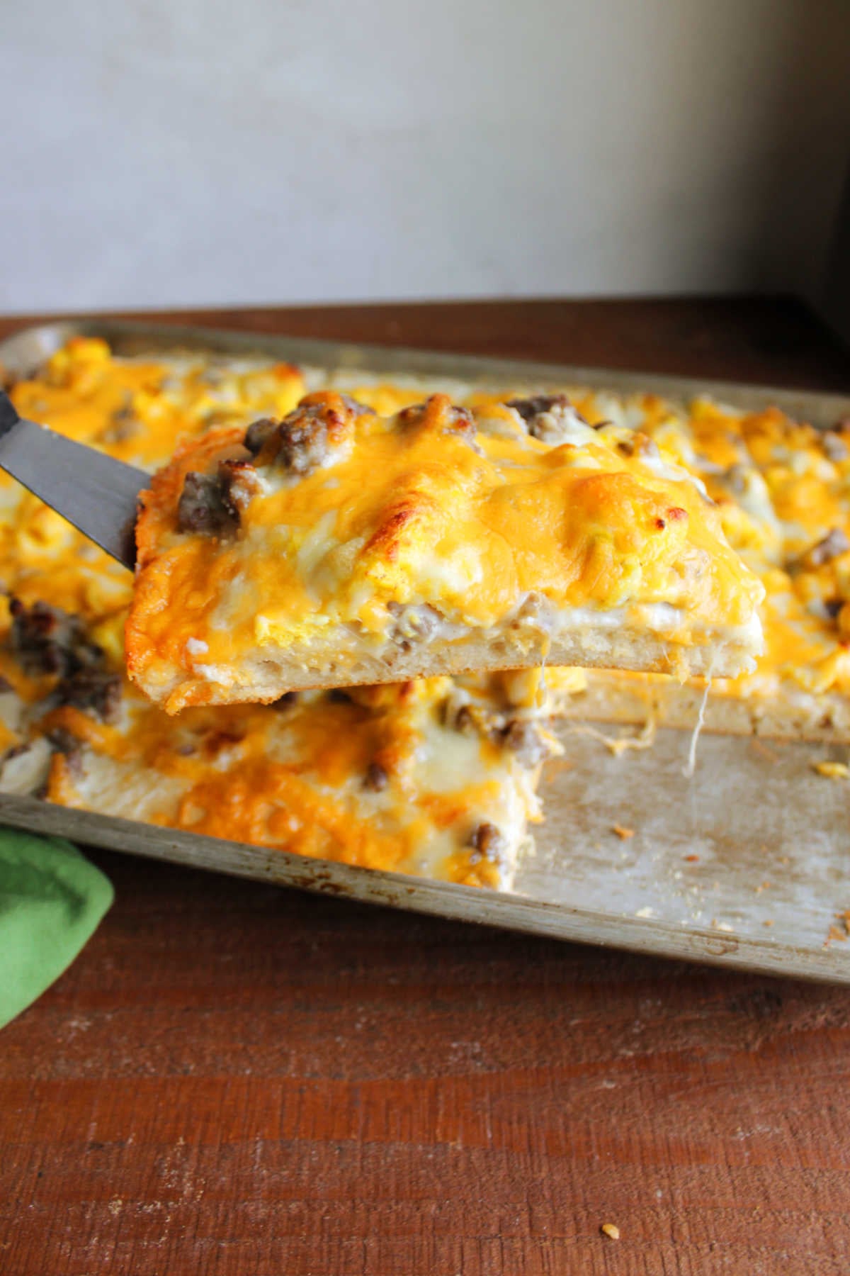Spatula lifting a large rectangular slice of breakfast pizza out of the sheet pan showing the crust topped with gravy, sausage, eggs, and cheese. 
