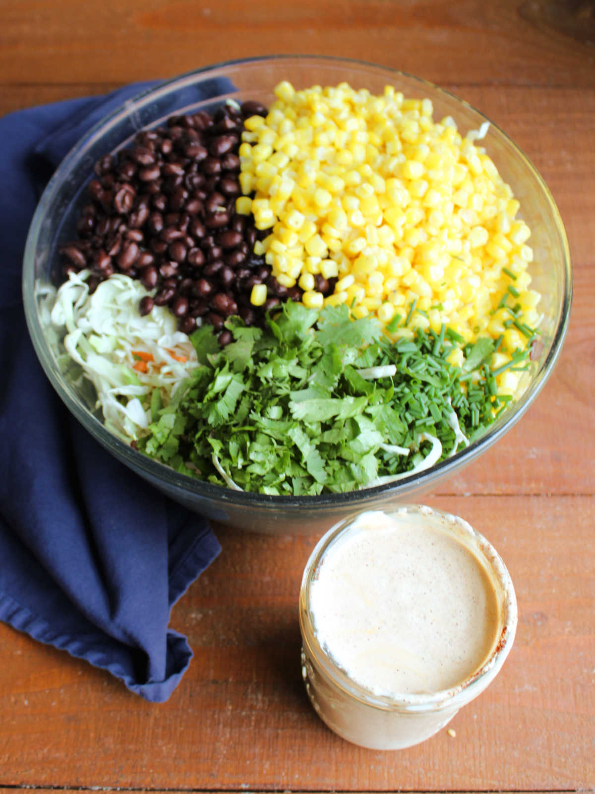 Large mixing bowl filled with coleslaw mix, corn, black beans, cilantro, and chives next to a jar of homemade creamy chili lime coleslaw dressing, ready to be mixed together. 