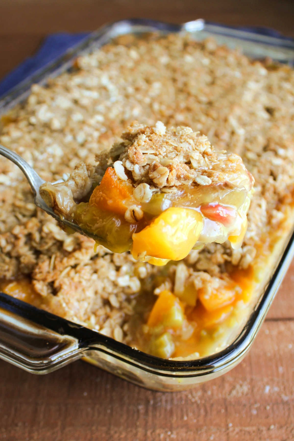 Serving spoon lifting a scoop of freshly baked peach rhubarb crisp with brown sugar oat topping out of the baking dish, ready to serve. 