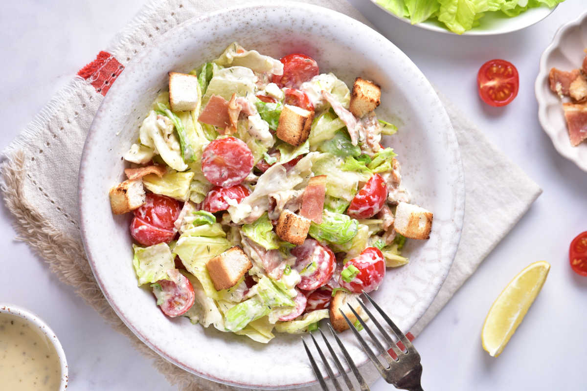 Bowl of BLT salad with extra dressing, lemon wedges, tomatoes, and bacon nearby. 